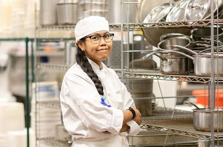 A young indigenous woman is wearing a chef's uniform and glasses and is standing in a kitchen in front of a shelf of pots and pans with her arms crossed.