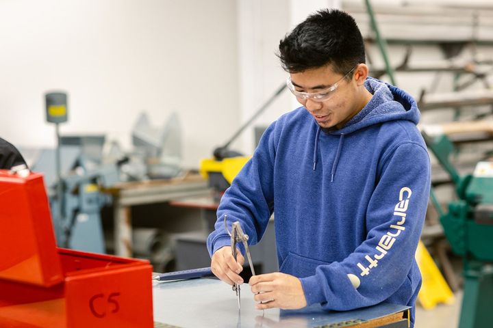 A student is using a tool in a Trades workshop. He is wearing a hoodie and protective glasses.