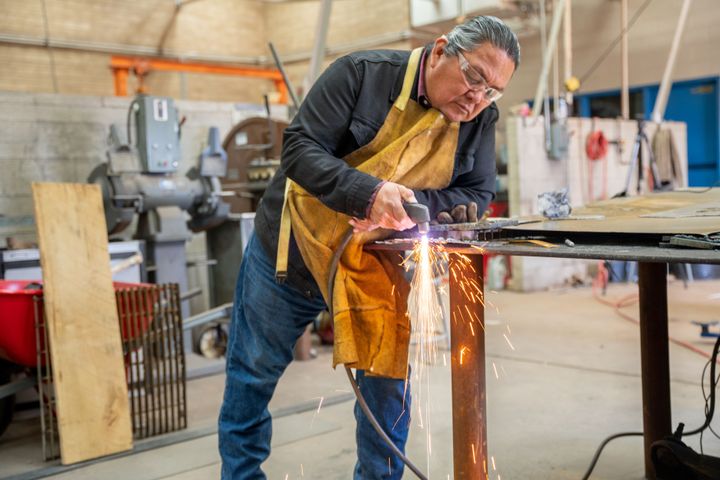 An older indigenous man is working in a wood shop wearing protective eyewear and an apron as he uses a power tool to cut wood. 
