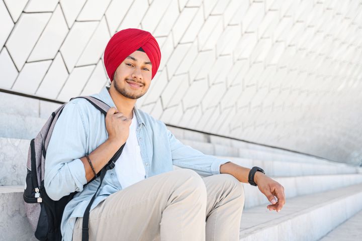 A young Sikh male is sitting on cement steps holding a backpack and smiling at the camera.