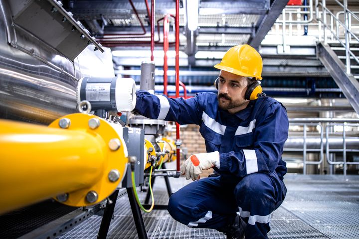 A male Trades student is wearing a yellow hard hat and protective ear and eye wear as well as a jumpsuit and gloves as he works on mechanical work.