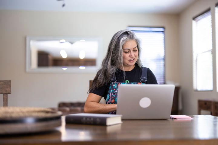 A woman with long grey hair is wearing overalls and sitting at a desk with a laptop and reading.
