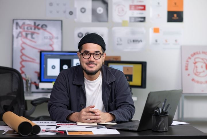 A male professional graphic designer sits as a desk wearing a toque and a round glasses await framed art and digital screens around him.