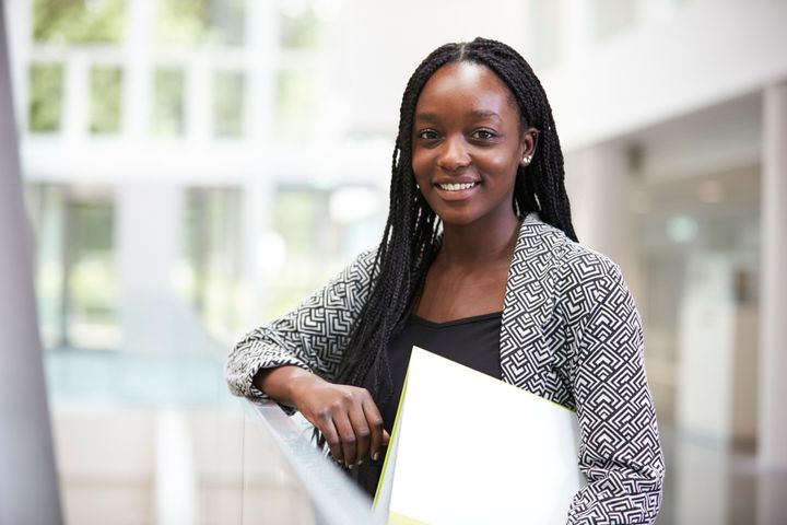 A female student is holding a file and posing in an institution against a railing and smiling at the camera.